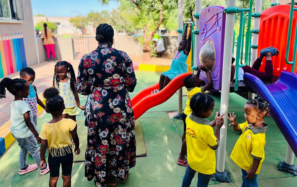 A teacher and children in yellow Zonke Izizwe uniforms on the preschool playground